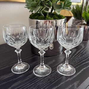 May include: Three clear crystal wine glasses with intricate cut patterns. The glasses have a rounded bowl shape, a slender stem, and a circular base. They are arranged on a dark wooden surface, with a silver planter and greenery in the background.