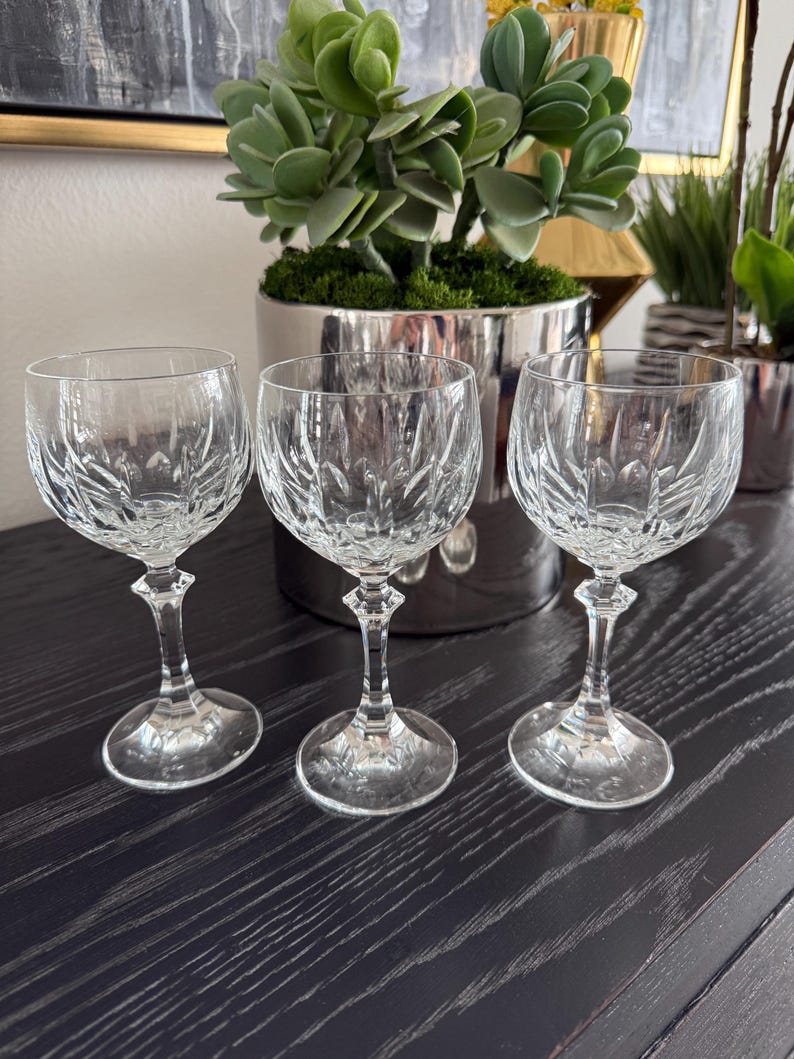 May include: Three clear crystal wine glasses with intricate cut patterns. The glasses have a rounded bowl shape, a slender stem, and a circular base. They are arranged on a dark wooden surface, with a silver planter and greenery in the background.