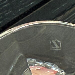 May include: A close-up of a silver-colored object with a rounded edge. The object has a reflective surface and a small, square-shaped marking with a stylized letter "S" inside. The background is dark.