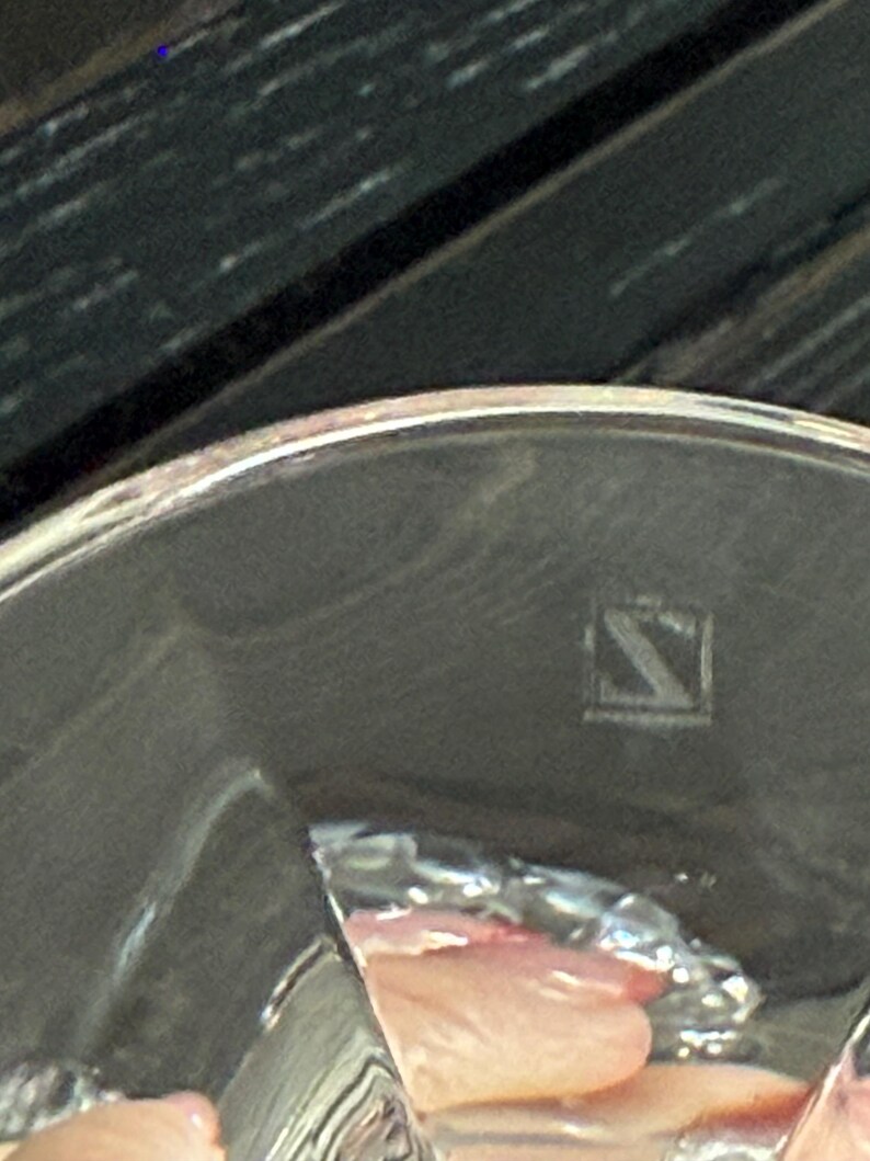 May include: A close-up of a silver-colored object with a rounded edge. The object has a reflective surface and a small, square-shaped marking with a stylized letter "S" inside. The background is dark.