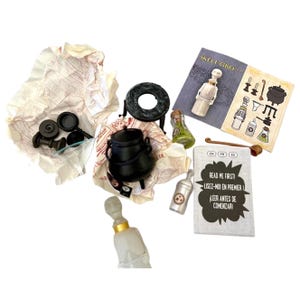 May include: A black cauldron, a small black stove, a glass bottle with a gold lid, and a small glass bottle with a green liquid. There are also two small black pans and a small black spoon. The image includes the text "READ ME FIRST! LISEZ-MOI EN PREMIER! LEER ANTES DE COMENZARI" in English, French, and Spanish.