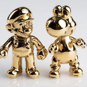 May include: Two gold-coloured figurines of video game characters, Mario and Yoshi, stand side-by-side. Mario wears a cap and overalls. Yoshi has a large head and a saddle-like structure on its back. Both figures have a shiny, reflective surface.