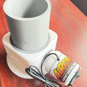 Puede incluir: Un dispositivo de fundición cilíndrico impreso en 3D de color blanco con un inserto de tubo vertical gris. Un pequeño componente de motor blanco etiquetado AIRCAS PERFECT CASTING EVERY está unido al lateral con un cable negro enrollado.