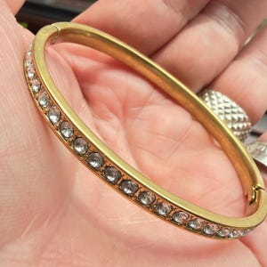 Puede incluir: Un brazalete rígido articulado de tono dorado que presenta una hilera de cristales transparentes de corte redondo incrustados en la superficie exterior. La pulsera tiene una banda de metal lisa con acabado mate y un cierre visible en el lateral.