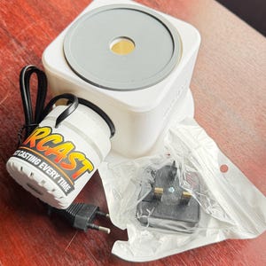 Peut inclure: Un appareil carré blanc avec un dessus circulaire gris et un trou central, relié à un composant cylindrique blanc portant l'inscription "ORCAST PERFECT CASTING EVERY TIME" avec un cordon d'alimentation noir et une prise électrique noire séparée.