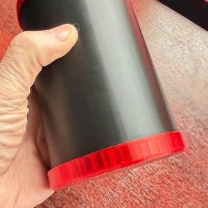 May include: A cylindrical container featuring a matte black body and bright red caps on both the top and bottom ends. The structure is smooth and uniform, with the red caps displaying a subtle vertical ribbed texture along their outer edges.