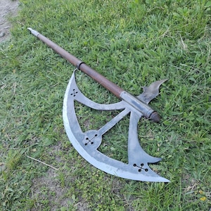 May include: A medieval poleaxe with a long wooden shaft and a large, curved steel blade. The blade features decorative cross-shaped cutouts. The axe is designed for combat and is lying on a grassy surface.