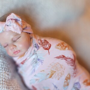 May include: A baby sleeping in a pink swaddle with a feather print. The baby is wearing a pink knotted headband.