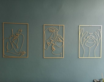 May include: Three gold metal wall sculptures featuring abstract line drawings of faces. The sculptures are framed in a thin gold metal frame.