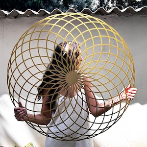 以下が含まれることがあります： 格子状のデザインが施された、大きな金色の幾何学的な球体が人の前に掲げられています。 球体は細い金属ストリップでできており、三次元効果を生み出しています。 背景には白い壁、緑、建物の様子が映っています。