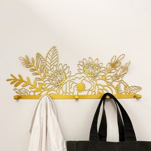 Op de afbeelding: Gouden metalen wandbevestigde kapstok met een bloemenmotief. De kapstok heeft vijf haken en is versierd met een uitsnijding van bloemen en bladeren. Een wit overhemd en een zwarte tote bag hangen aan de haken.
