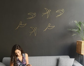 May include: A woman sits on a gray and white checkered sofa reading a magazine. The wall behind her is dark gray and decorated with gold wire bird sculptures.