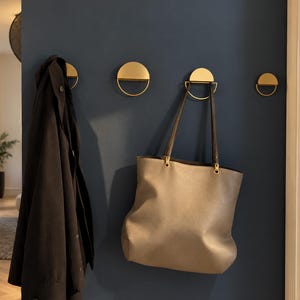 Puede incluir: Cuatro ganchos de pared dorados, cada uno con una parte superior de semicírculo sólido y una base de bucle de alambre semicircular delgado. Los ganchos están montados en una pared azul oscuro, con una chaqueta oscura en el primero y un bolso beige en el tercero.