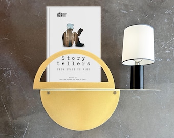 May include: A gold metal shelf with a rounded front edge holds a white book titled "Story Tellers: From Stage to Page" and a white lamp with a black base. The book features a silhouette of a person sitting and reading.