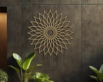 May include: A gold metal wall decor in the shape of a large flower with intricate details. The decor is mounted on a gray concrete wall. There are three potted plants in front of the wall.