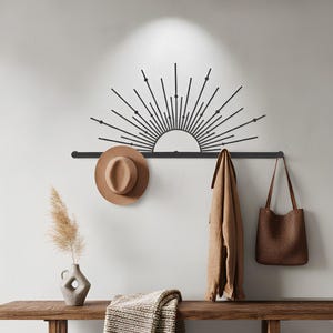 Può includere: Un attaccapanni da parete in metallo nero con un design a raggiera, che tiene un cappello marrone, un cappotto beige e una borsa tote marrone. Una panca in legno sottostante contiene una coperta piegata e un vaso con piante essiccate.