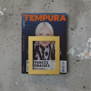 May include: A gold picture frame with a photograph, placed on a magazine titled "TEMPURA." The magazine features a woman with blonde hair and the text "NOUVEAU." The frame is square and the magazine is black and blue.