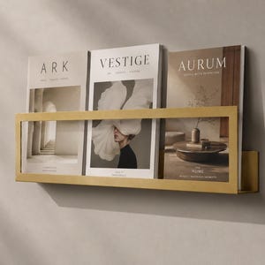 May include: A rectangular gold-toned metal wall-mounted display rack holding three magazines titled ARK, VESTIGE, and AURUM. The rack features a simple frame design that keeps the magazines upright and visible against a neutral wall surface.