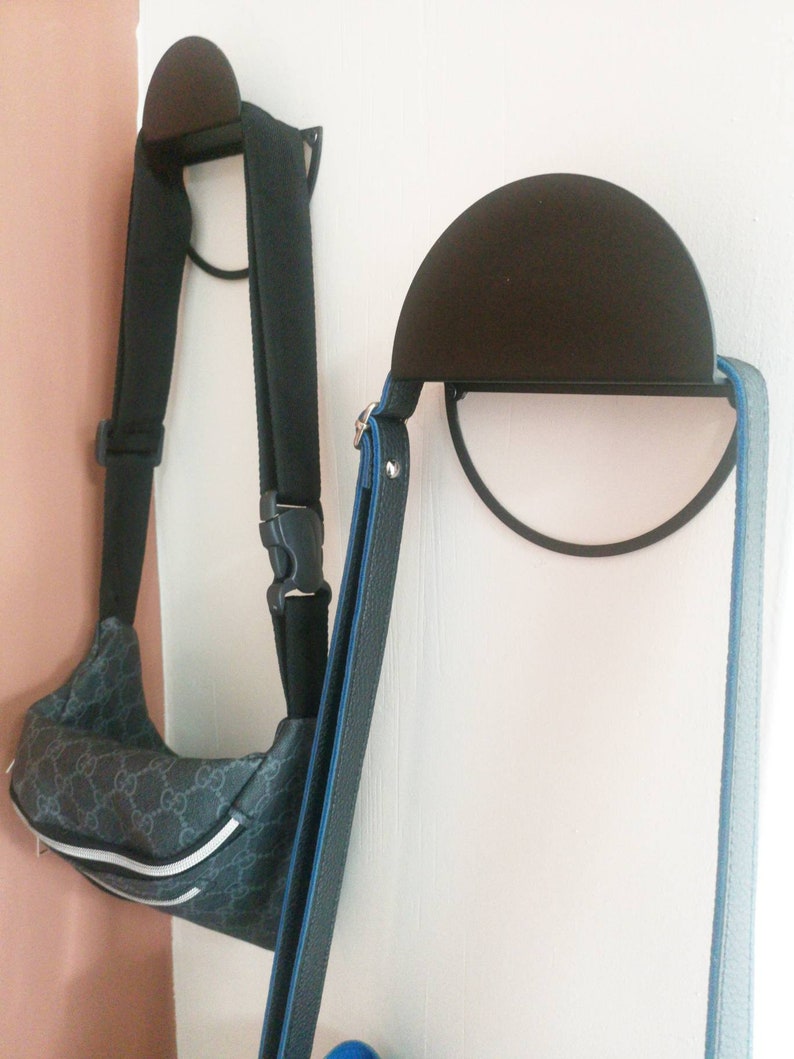 Peut inclure: Deux crochets muraux en m&eacute;tal noir avec des sommets arrondis. Un crochet a un sac &agrave; motifs noir et gris suspendu. L'autre crochet a une sangle bleue et noire suspendue.