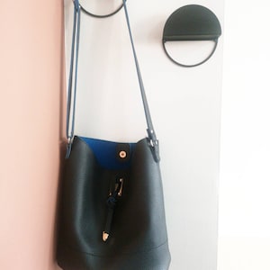 Peut inclure: Un sac seau en cuir noir avec une doublure bleue et une boucle dor&eacute;e accroch&eacute;e &agrave; un crochet en m&eacute;tal noir.