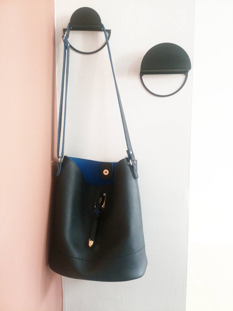 Peut inclure: Un sac seau en cuir noir avec une doublure bleue et une boucle dor&eacute;e accroch&eacute;e &agrave; un crochet en m&eacute;tal noir.