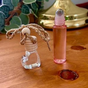 May include: A clear, heart-shaped glass bottle with a wooden top and a rope hanger, next to a pink roller bottle. The bottles are on a wooden surface. The roller bottle is filled with a pink liquid.