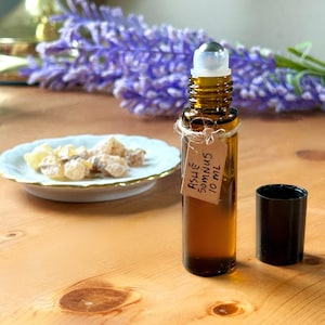 May include: A brown glass roll-on bottle with a silver rollerball and a black cap. A small tag tied to the bottle reads "ASHÉ Somnus 10 ml." A small white plate with gold trim holds several light-colored, irregularly shaped pieces. Purple flowers are in the background.