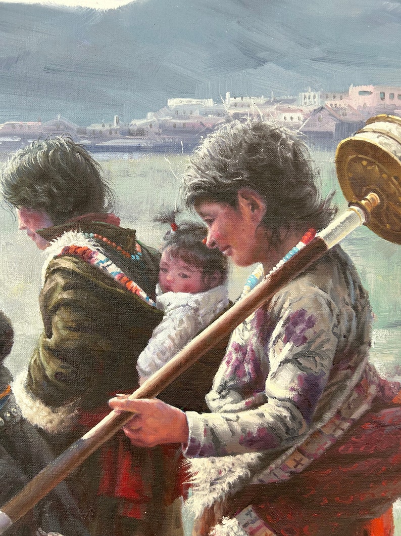 May include: A woman in a floral patterned shirt carries a child on her back while holding a long wooden stick with a metal prayer wheel attached. The woman is wearing a red and white scarf and a white shawl. The child is wearing a white shirt and a white hat.