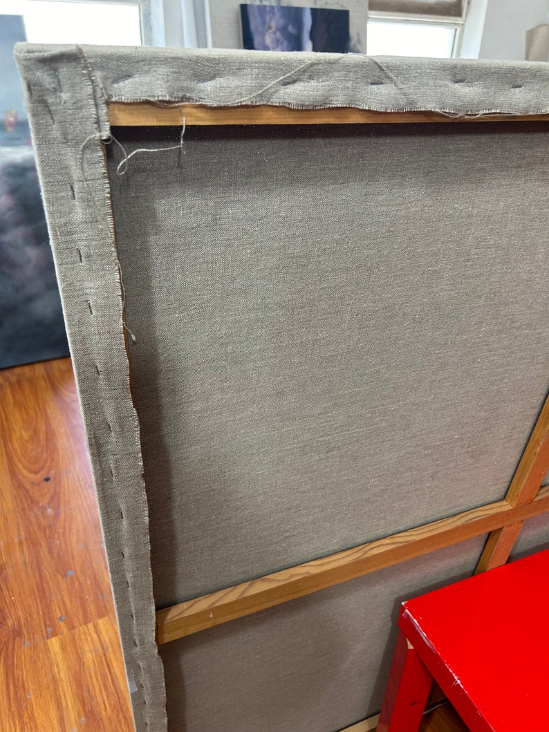 May include: The back of a canvas painting with a wooden frame. The canvas is stretched tightly over the frame and is secured with staples. The frame is made of natural wood and has a light brown color.