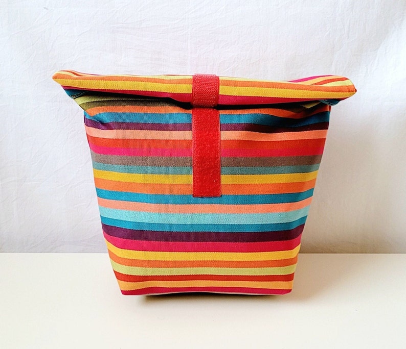 Eine bunt gestreifte Lunchbag mit Innenfutter aus abwaschbarer beschichteter Baumwolle rot weiß gepunktet.Die Lunchbag ist 30 cm hoch, 28 cm breit und 10 cm tief.Zum  Auf und Zumachen ist ein rotes Klettband aufgenäht.