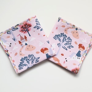 Könnte beinhalten: Zwei rosa Stoffquadrate mit einem bunten Druck eines Schlosses, Bäumen und einem Einhorn. Der Stoff hat einen rosa Hintergrund mit einem bunten bestickten Rand.