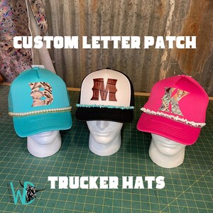 May include: Three trucker hats with custom letter patches. The hats are turquoise, black, and pink. The letters are "B", "M", and "K".