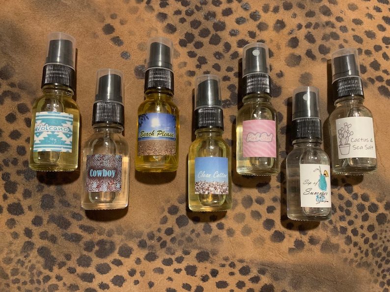 May include: Eight small glass bottles with spray tops and labels. The labels have various designs and text, including "Volcano", "Cowboy", "Beach Please", "Clean Cotton", "Pink Salt", "Sip of Summer", and "Cactus & Sea Salt".