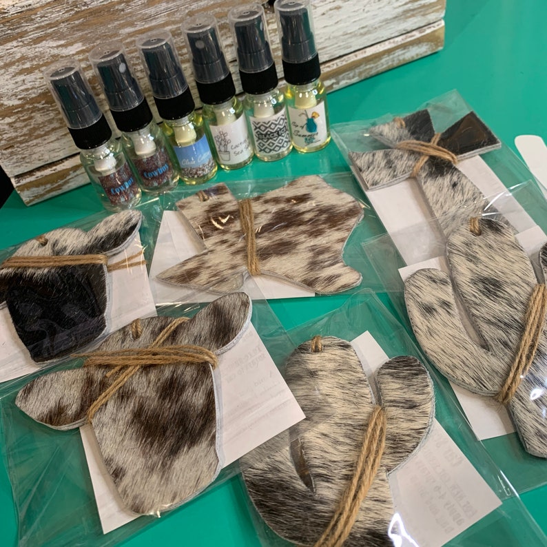 Puede incluir: Seis peque&ntilde;as botellas de aceite esencial con etiquetas que dicen "Cowboy", "Lychee", "Summer", "Coffee", "Vanilla" y "Sage". Las botellas est&aacute;n dispuestas en una fila sobre una superficie de madera blanca. Debajo de las botellas hay seis ambientadores de coche con estampado de piel de vaca en varias formas, incluyendo un cactus, una herradura y un estado de Texas.