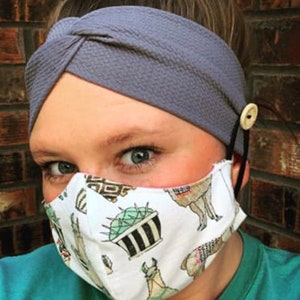 May include: A woman wearing a gray headband with a knot on top and a white face mask with a llama and cactus print. The mask has a button on each side for attaching ear loops.