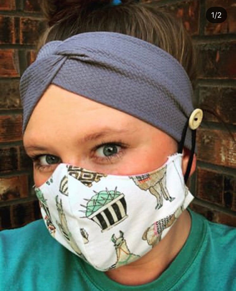 May include: A woman wearing a gray headband with a knot on top and a white face mask with a llama and cactus print. The mask has a button on each side for attaching ear loops.