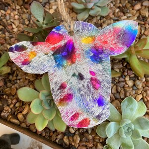 May include: A colorful cowhide ornament with a white background and iridescent pink, blue, green, and yellow accents. The ornament is hanging from a twine string.