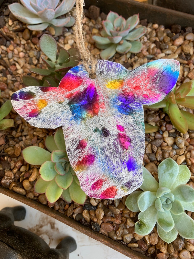 May include: A colorful cowhide ornament with a white background and iridescent pink, blue, green, and yellow accents. The ornament is hanging from a twine string.