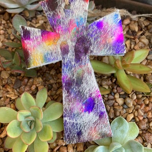 May include: A white leather cross with a colorful, iridescent, tie-dye pattern. The cross is hanging from a string and is surrounded by small green succulents and brown rocks.