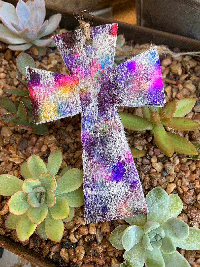May include: A white leather cross with a colorful, iridescent, tie-dye pattern. The cross is hanging from a string and is surrounded by small green succulents and brown rocks.