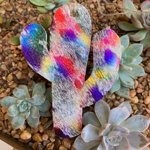 May include: A colorful cactus-shaped ornament made of leather with a white background and iridescent rainbow accents. The ornament is hanging from a twine string.