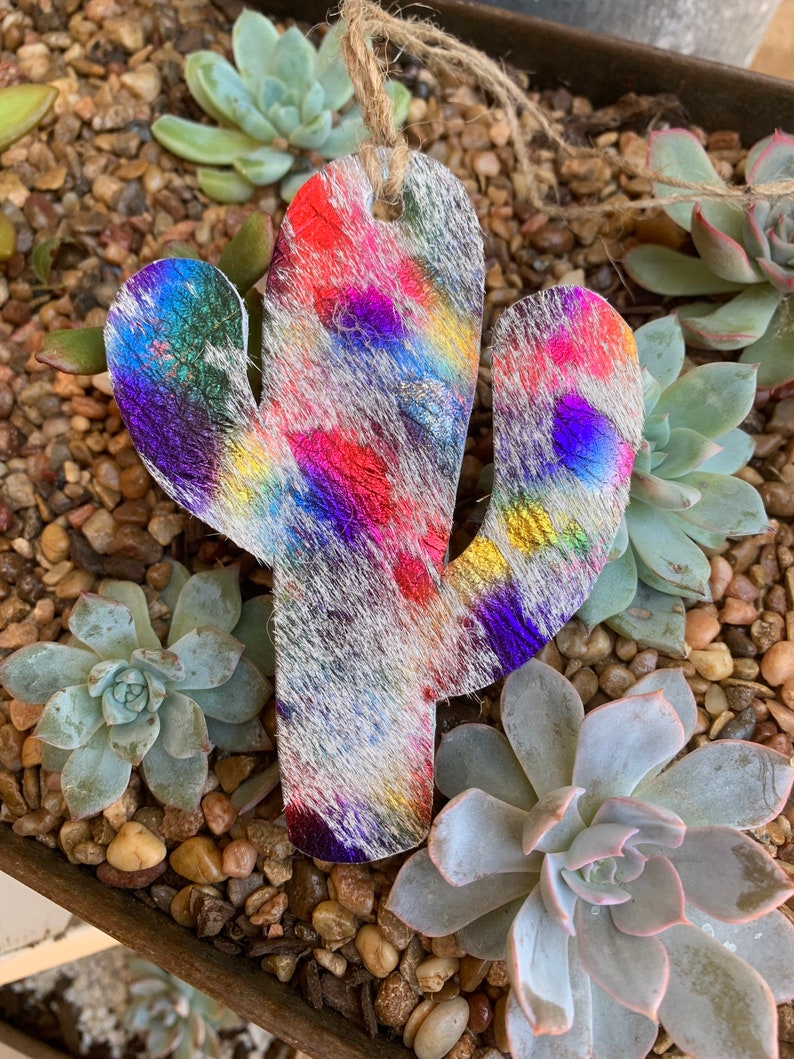 May include: A colorful cactus-shaped ornament made of leather with a white background and iridescent rainbow accents. The ornament is hanging from a twine string.