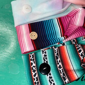 May include: Three colorful headbands with a knotted design. Each headband has a different pattern and a button. The top headband is a light blue and pink tie-dye pattern with a white button. The middle headband is a pink, blue, and black striped pattern with a wooden button that says "Handmade with Love". The bottom headband is a green, orange, and black striped pattern with a black button.