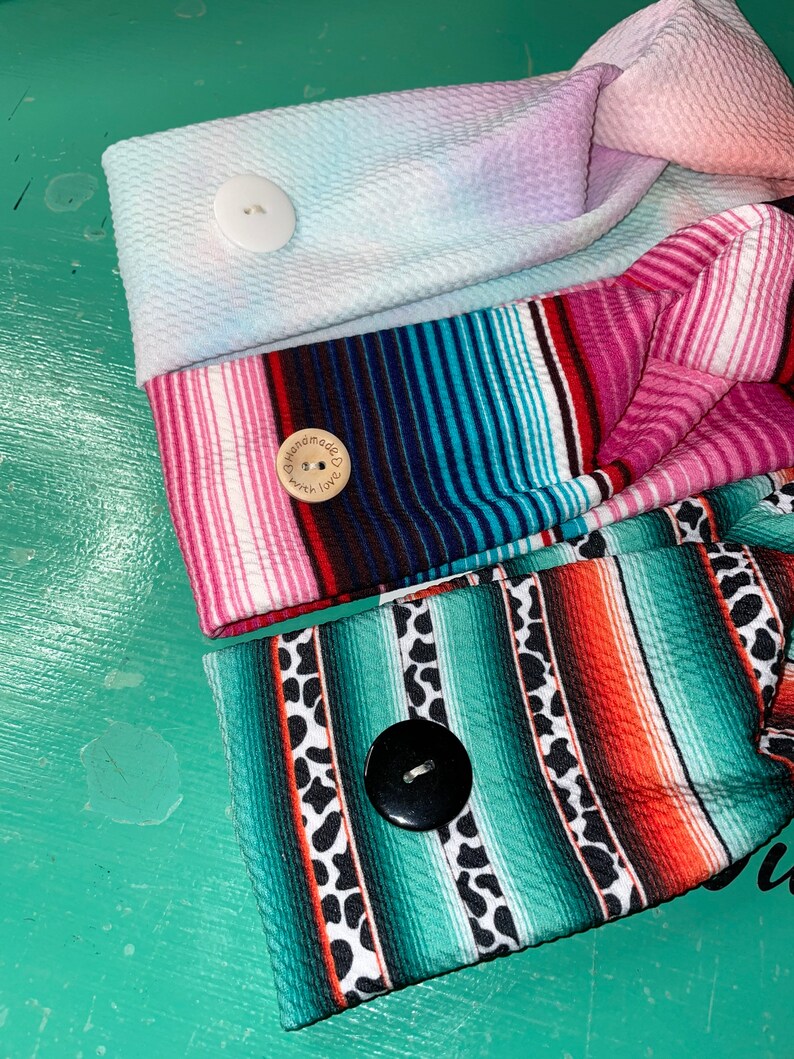 May include: Three colorful headbands with a knotted design. Each headband has a different pattern and a button. The top headband is a light blue and pink tie-dye pattern with a white button. The middle headband is a pink, blue, and black striped pattern with a wooden button that says "Handmade with Love". The bottom headband is a green, orange, and black striped pattern with a black button.