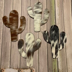 Puede incluir: Cuatro formas de cactus recortadas de piel de vaca con diferentes patrones de colores. Las formas de cactus son marrones, negras y blancas.