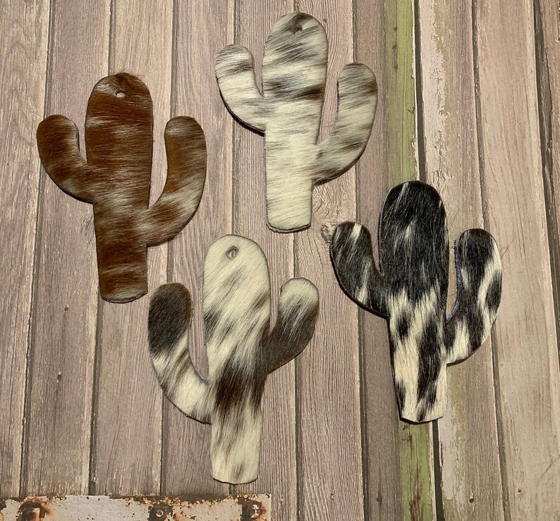 Puede incluir: Cuatro formas de cactus recortadas de piel de vaca con diferentes patrones de colores. Las formas de cactus son marrones, negras y blancas.