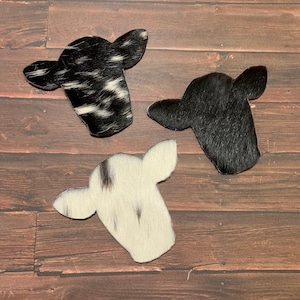Puede incluir: Tres parches de piel de vaca en forma de cabeza de vaca. Los parches son blanco y negro, negro y blanco con manchas negras.