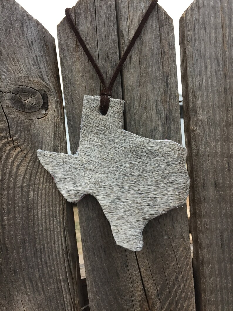 Puede incluir: Un adorno de cuero blanco con forma de Texas que cuelga de un cord&oacute;n de cuero marr&oacute;n en una valla de madera.