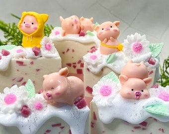 May include: Four pink and white layered soap bars with a pink and white swirl design. Each bar is topped with a small pink pig figurine and white flowers. One bar has a yellow pig figurine wearing a yellow raincoat.