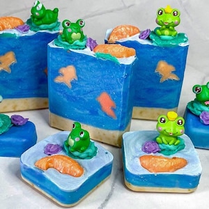 May include: A set of six handmade soap bars featuring a blue and white design with orange accents. Each bar has a small green frog figurine on top. The soap bars are arranged in a row on a white surface.
